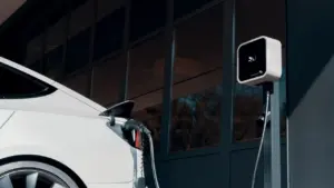 Balts elektroauto, kas pieslēgts lādēšanas stacijai modernā pilsētvidē ārpus telpām