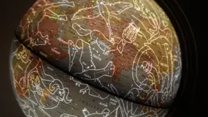 Tuvplānā redzams globuss ar attēlotām zodiaka zīmēm un zvaigznājiem tumšā fonā.