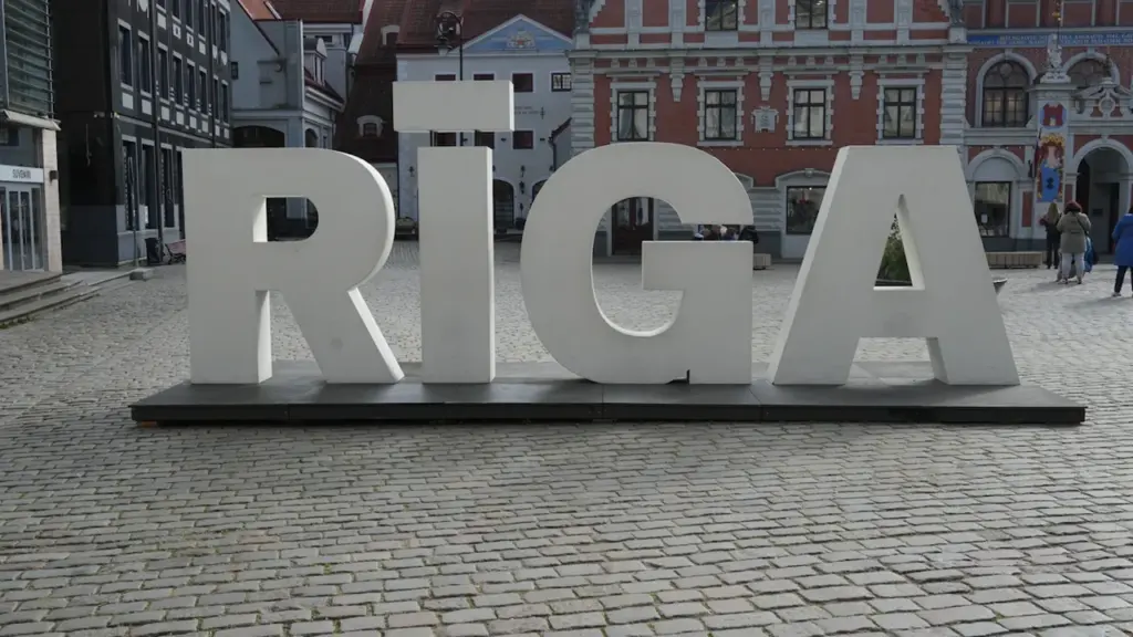 Liels, balts uzraksts RĪGA Rīgas Rātslaukumā ar bruģakmens ielu un vēsturiskām ēkām.