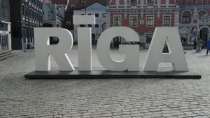 Liels, balts uzraksts RĪGA Rīgas Rātslaukumā ar bruģakmens ielu un vēsturiskām ēkām.